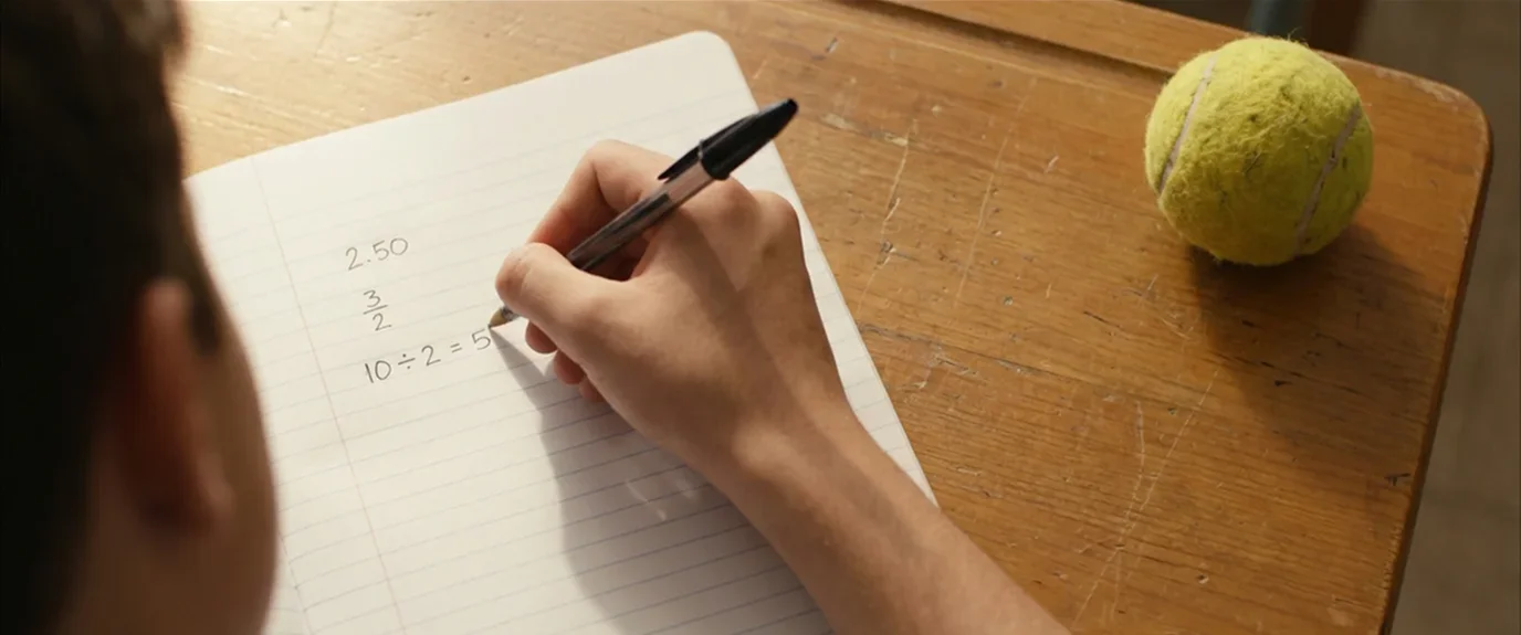 Primer plano de una mano escribiendo cálculos con bolígrafo en un cuaderno junto a una pelota de tenis
